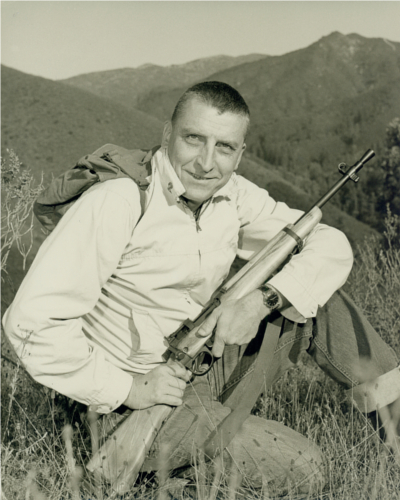 Con Sellers sitting in a field, holding a British Rifle .303 No. 5 Mk I, with a backdrop of mountains.