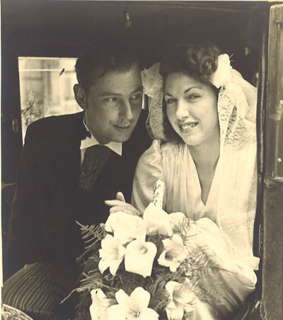 This black and white photograph shows Anny van Bommel and John Garrison on their wedding day in Belgium. The couple is sitting in a carriage with the bride in a wedding dress holding a bouquet of flowers. The groom is looking at the bride and smiling and is dressed in formal garb including a black jack and vest and striped pants.