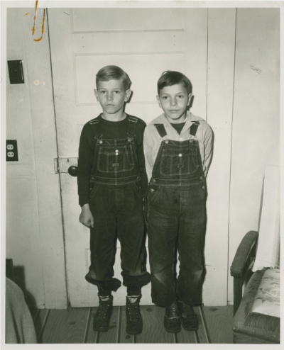 Two boys in overalls standing in front of a house door with a chair to the right of them.  