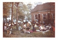 Photograph of a group of people sitting on a lawn under trees, behind a red brick building. Photo is of a meeting of the Council of Federated Organizations.