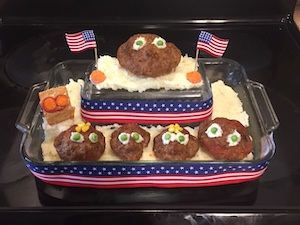Pan of mashed potatoes, with meat patties on top. Meat patties have peas for eyes. One patty is on another box with American flags on either side. Creation resembles a speaker and an audience