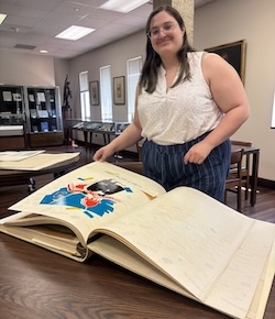Ella Lauderdale stands beside a large open archival scrapbook displayed on a table in a library or archives reading room. The oversized volume contains a colorful illustrated page featuring a bird and a photograph. Display cases and framed artwork are visible in the background.