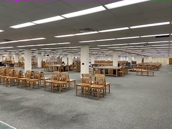 photo is a large room with green carpet. At the front of the photo is a row of chairs, back to back. Behind the chairs are white columns with wooden tables and chairs around the columns.