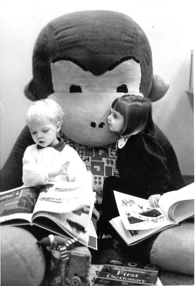 : A black and white photo of two young children reading on Big George’s lap. The little boy, Garrett Rabian, has light hair and is wearing a white sweater. The photograph catches him flipping excitedly through the large book in his lap. The little girl, Janice Rabian, has dark hair cut into a bob and bangs; she’s wearing a black dress with a white lace collar. In the photo, she is hugging Big George’s neck, her cheek against his, while she holds a copy of Curious George in her lap. 