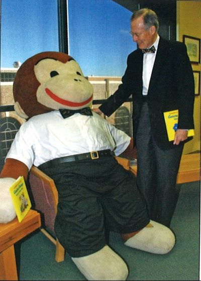 A photo of Dr. Aubrey Lucas standing with Big George, holding a copy of one of the Curious George books. Big George is wearing black slacks and a plain white dress shirt with a black bow tie. Dr. Lucas stands next to George, smiling down at him with a hand on his shoulder. Dr. Lucas matches George’s outfit with an added black blazer. 
