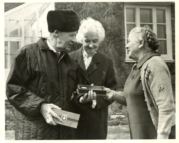 Black and white photograph of E. H. Shepard receiving the USM Medallion from Dr. Lena Y. de Grummond while Mrs. Norah Shepard observes. 