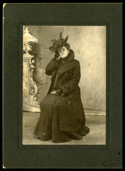 Formal studio photo of Katherine Brown seated in overcoat, gloves, and hat with feathers. She has one handheld to her head. There is a decoration of a Roman column in the background.