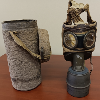 The image is of a gas mask and a paper mache storage cannister. Both are approximately 11” high are a dark khaki color. The cannister is 5 inches in diameter. It has a rough texture and a lid affixed by cloth straps. The gas mask has a minimal design consisting of goggles and a cloth mask affixed to a metal base. The letters DUCL are printed on the cloth mask and 10P50 and P.T are printed on the straps. A pipe connects the metal base to a metal filter cartridge that is imprinted on the bottom with the model number, C.P. 1935 M. Marked on the bottom of the cartridge in white ink is C.C.F. 45-1 and two indecipherable letters enclosed in a circle. The plastic eye coverings are yellowed, shriveled, and torn with age. 